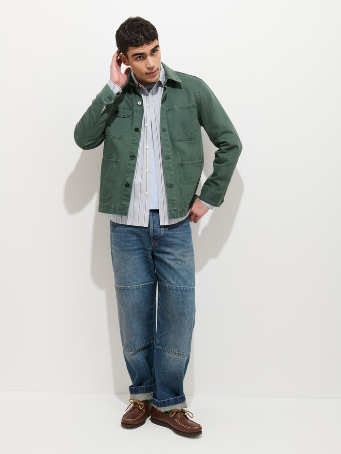 A man stands against a white background, wearing the Garment Dyed Work Jacket In Recycled Denim over a striped shirt, blue jeans with knee patches, and brown boat shoes. He has short curly hair and touches his left ear while glancing to the side.