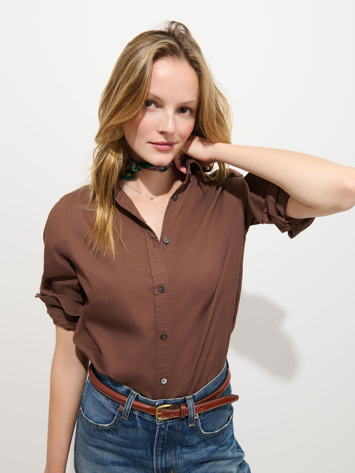 A woman with long blonde hair wears the Gracie Shirt in Poplin, a classic button-up brown shirt crafted from 100% cotton, paired with jeans and a brown belt. She stands against a plain white background with one arm raised, touching her neck.