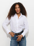 A person with curly hair stands against a white background, looking at the camera with their hands on their waist, wearing the Gracie Shirt in Poplin and blue jeans.