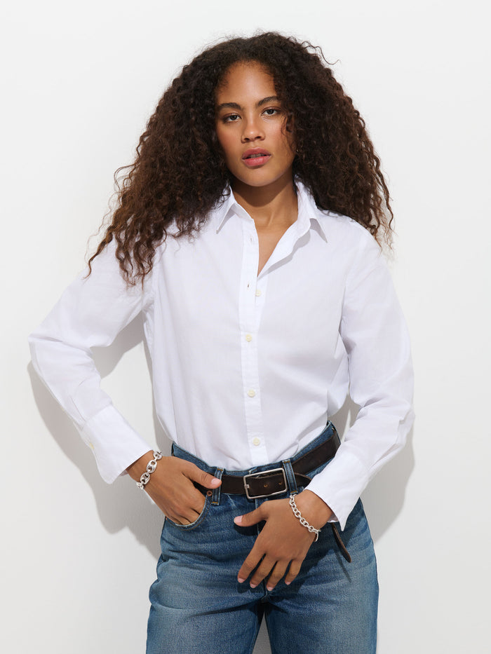A person with curly hair stands against a white background, looking at the camera with their hands on their waist, wearing the Gracie Shirt in Poplin and blue jeans.