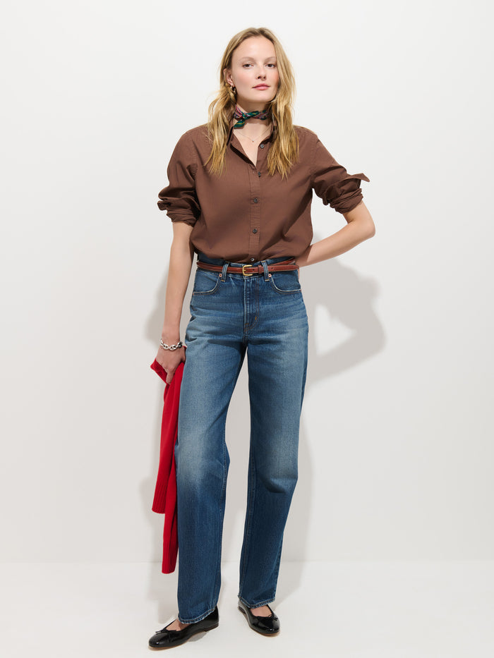 A woman stands against a white background wearing the Gracie Shirt in Poplin—a classic, button-up brown shirt—paired with blue jeans, black flats, and a neck scarf. She holds a red jacket in her left hand and has her right hand on her hip.