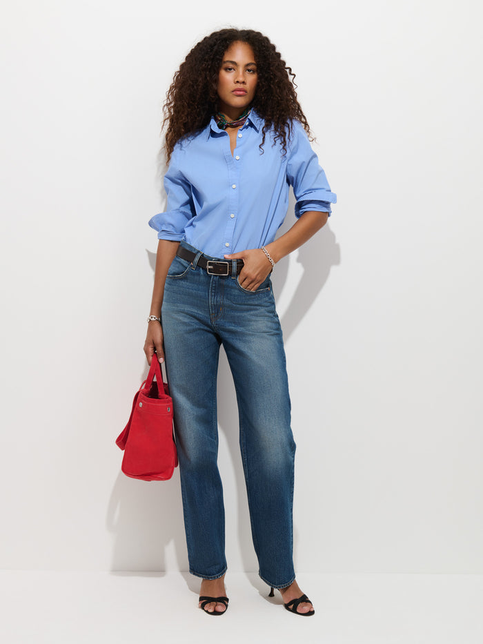 A woman stands against a white background wearing the Gracie Shirt in Poplin, a classic button-up light blue shirt crafted from 100% cotton. She pairs it with blue jeans, black sandals, and carries a red handbag. Her curly hair falls softly as she wears a necklace, with her sleeves stylishly rolled up.