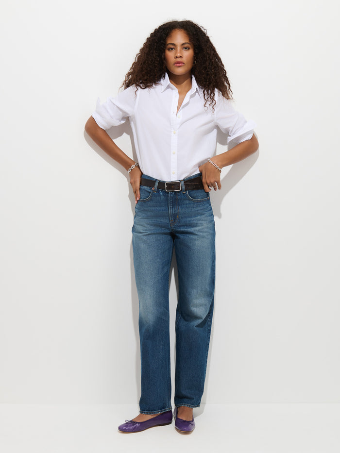 A woman with curly hair stands against a plain white background, hands on her hips, wearing the Gracie Shirt in Poplin, blue jeans, and purple flats.
