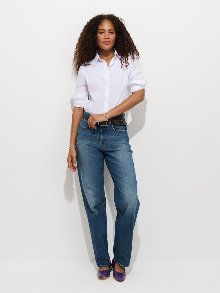 A woman with curly hair wearing the Gracie Shirt in Poplin, blue jeans, and purple shoes stands against a white background, looking at the camera with a relaxed pose.