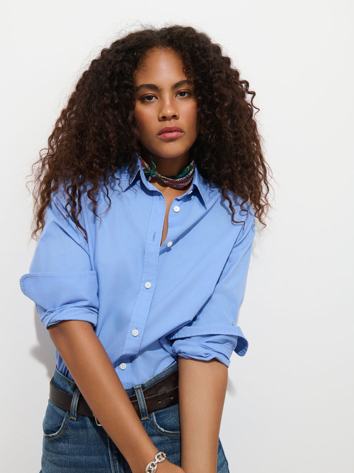 A woman with long curly hair wears the Gracie Shirt in Poplin in blue and jeans, standing against a white background with a neutral expression.