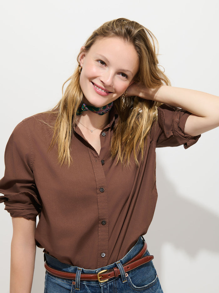 A smiling woman with blonde hair, wearing the Gracie Shirt in Poplin, blue jeans with a brown belt, and a floral scarf around her neck, stands against a plain white background with one hand behind her head.