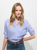 A woman with long blonde hair wearing the Gracie Striped Shirt in Poplin and jeans stands against a plain white background, posing with one hand on her hip and a slight smile.