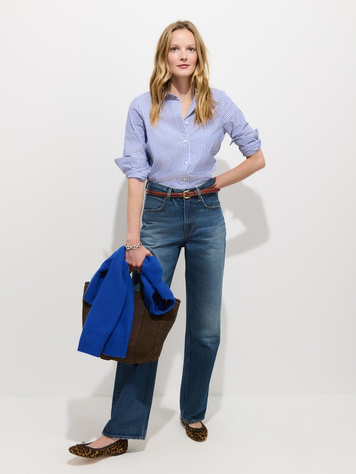 A woman stands against a white background wearing the Gracie Striped Shirt in Poplin, paired with jeans, leopard flats, and holding a blue sweater and brown tote bag. Her hair is loose, and one hand rests on her hip.