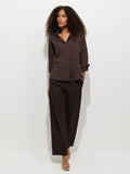 A woman with curly hair stands against a plain white background, looking slightly to the side, wearing a loose-fitting dark brown button-up shirt paired with the Hudson Pant in cotton twill.