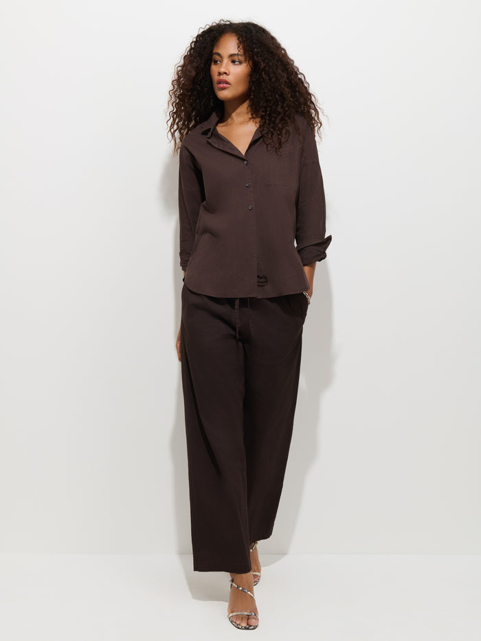 A woman with curly hair stands against a plain white background, looking slightly to the side, wearing a loose-fitting dark brown button-up shirt paired with the Hudson Pant in cotton twill.