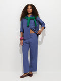 A woman with long curly hair stands against a white background, wearing the Hudson Pant In Cotton Twill—a pair of blue wide leg pants—paired with a matching blue button-up shirt. She has a green sweater draped over her shoulders, accessorizes with pink bracelets, and completes the look with brown flats.