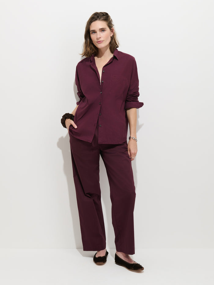 A woman stands against a white background wearing the Hudson Pant In Cotton Twill, styled in a loose, wide-leg fit with a matching maroon button-up shirt and black flats. One hand is in her pocket as she poses with a relaxed, confident expression.