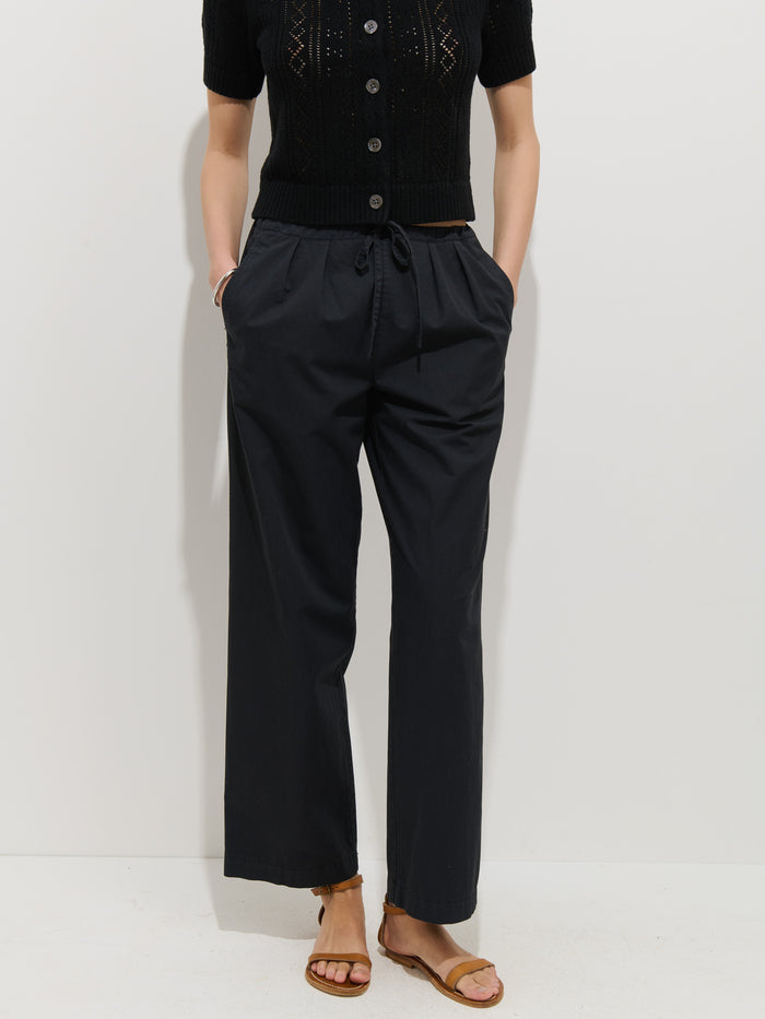 A person wearing a black short-sleeve button-up top, the Hudson Pant In Cotton Twill with a drawstring waist, and brown flat sandals stands against a plain white background with hands in their pockets.