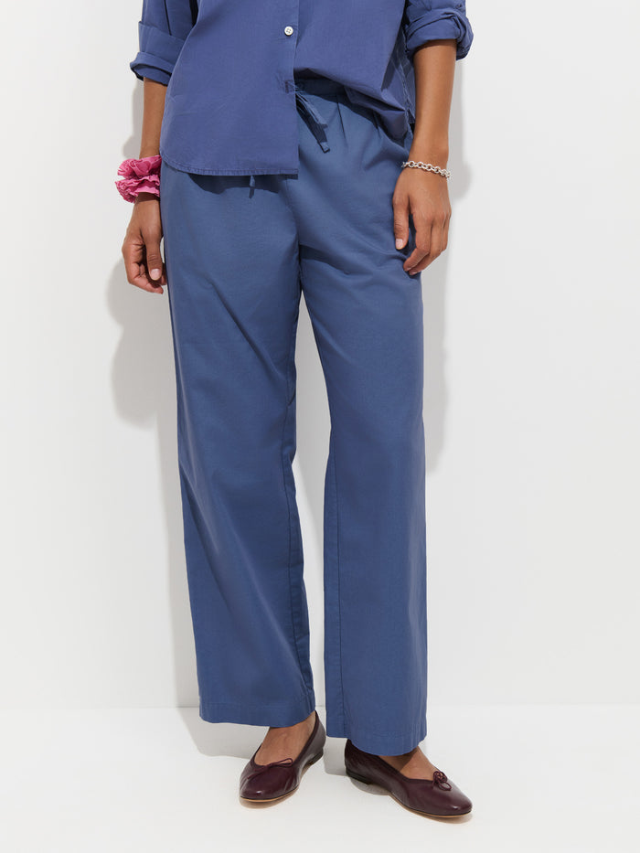 A person stands against a white background, wearing the Hudson Pant in Cotton Twill, styled in a loose blue wide-leg silhouette paired with a matching blue shirt. They have a pink scrunchie on one wrist and are wearing brown loafers. Their head is not visible in the image.