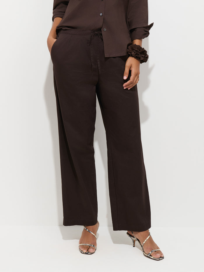 A person wearing the Hudson Pant In Cotton Twill in a loose, dark brown wide-leg style and a matching shirt stands against a white wall. One hand is in a pocket, and they wear snake-patterned high-heeled sandals.