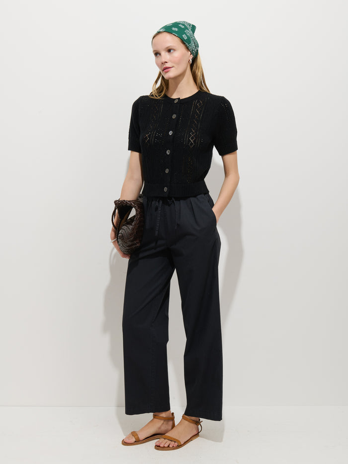 A woman stands against a plain white background wearing a green headscarf, a black short-sleeve button-up knit top, Hudson Pant in Cotton Twill, brown sandals, and holding a brown clutch.