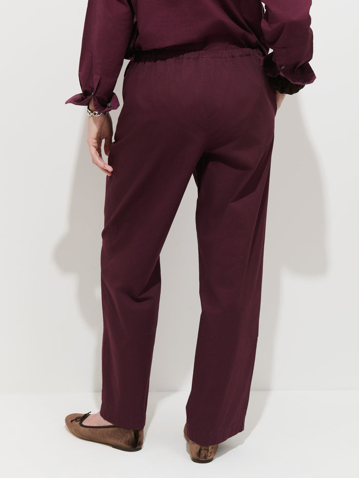 Wearing the Hudson Pant In Cotton Twill in burgundy, a person stands facing a white wall with only their lower back, wide leg pants, and brown flat shoes visible.