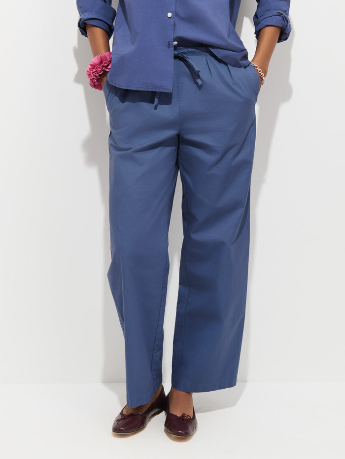 Person wearing loose blue Hudson Pant In Cotton Twill, a matching blue button-up shirt, maroon loafers, and a pink scrunchie on their wrist, standing against a white background. Their face is not visible.