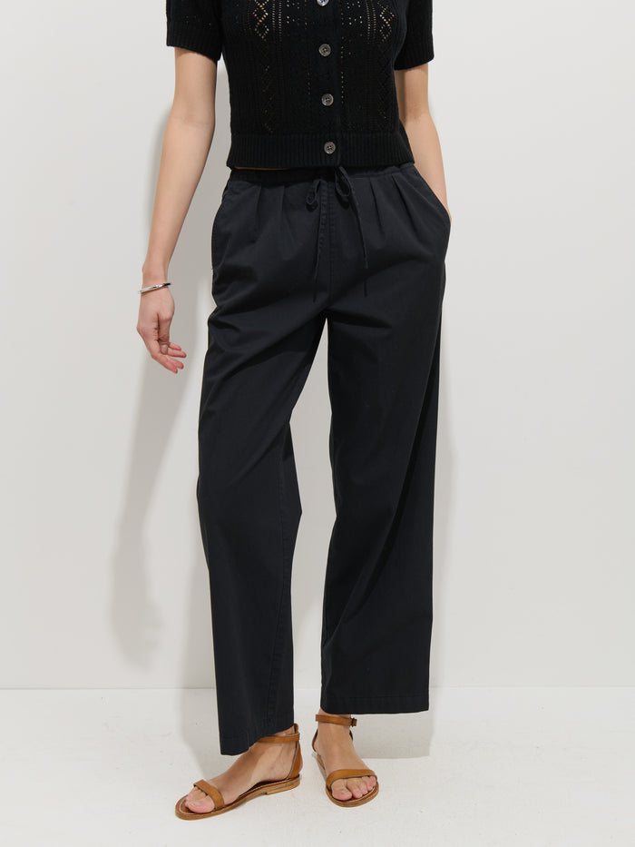 Wearing the Hudson Pant In Cotton Twill with a short-sleeve black button-up top and brown sandals, a person stands against a plain white background, visible from shoulders down.