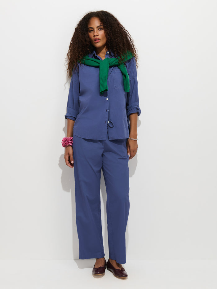 A woman with curly hair stands against a white wall wearing the Hudson Pant In Cotton Twill—a blue, wide-leg style—paired with a matching button-up shirt and a green sweater draped over her shoulders. She accessorizes the look with brown shoes and pink bracelets on her wrist.