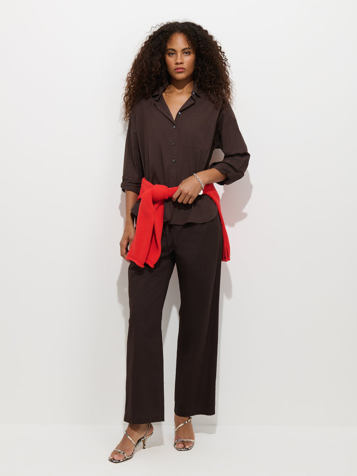 A woman with curly hair stands against a plain white background, wearing a dark brown button-up shirt and the Hudson Pant In Cotton Twill in a matching shade. She has a red sweater tied around her waist and completes the look with silver heels.