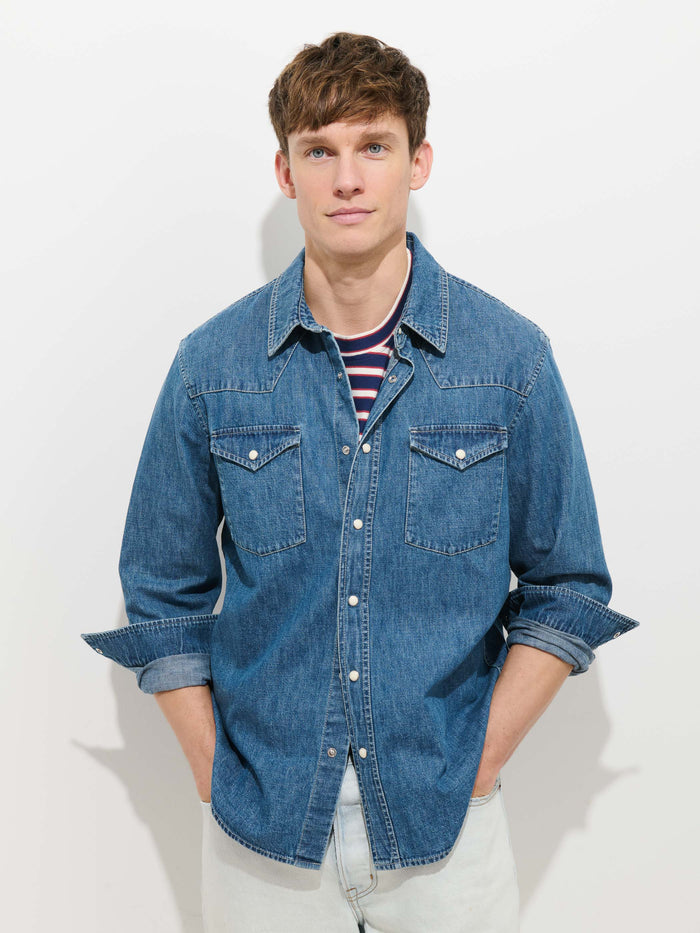 A young man stands against a white background, wearing the Jackson Pocket Shirt In Denim over a striped t-shirt, hands in his pockets and sleeves rolled up.