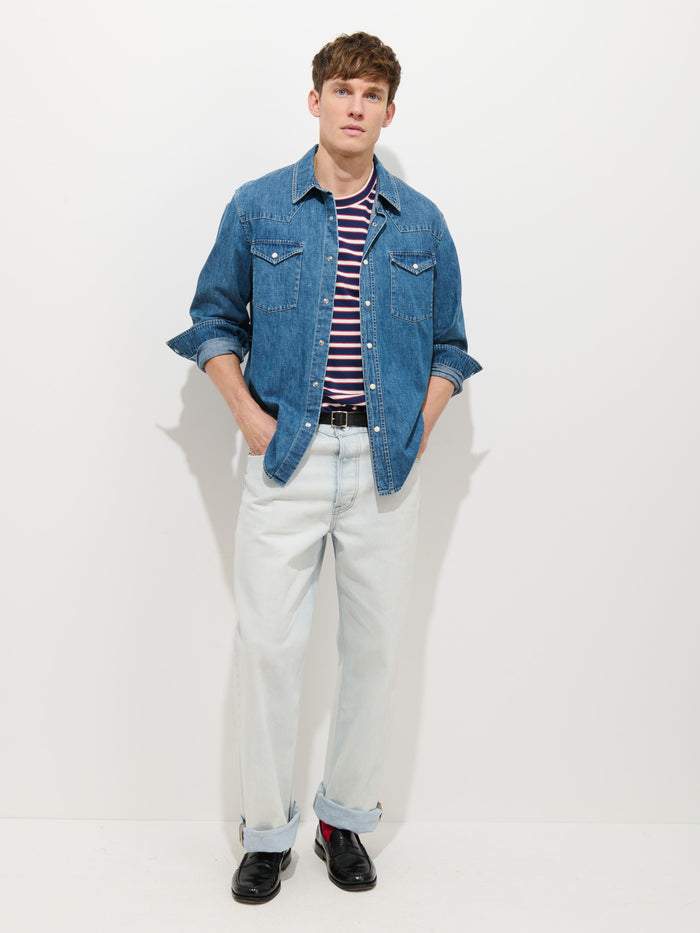A person stands against a plain white background wearing the Jackson Pocket Shirt in Denim over a red and white striped t-shirt, with light blue cuffed jeans, a black belt, and black shoes. Their hands are tucked into their pockets.