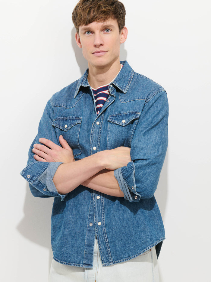 A young man stands against a white background, arms crossed, wearing the Jackson Pocket Shirt In Denim over a striped T-shirt with light-colored pants, and looks at the camera with a neutral expression.