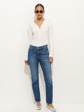 A woman stands against a white wall wearing sunglasses, a white long-sleeve henley, Jane Mid Rise Straight Jean, and silver ballet flats. Her hair is tied back and her hands are in her pockets.