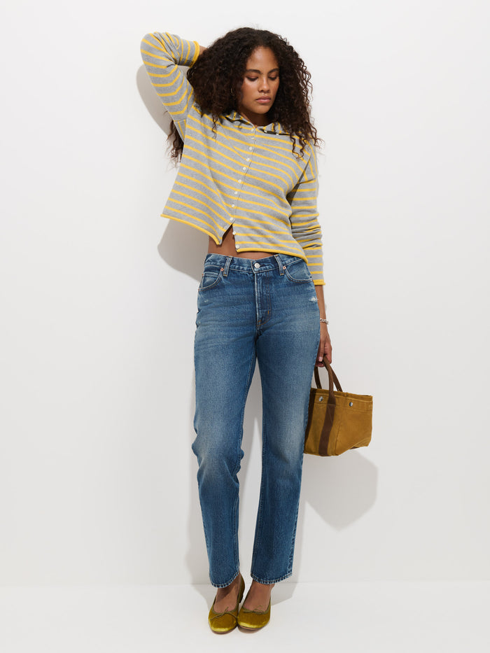 Woman with curly hair wears a yellow striped sweater, Jane Mid-Rise Straight Jean in 100% cotton denim, and yellow shoes, holding a brown handbag, standing against a plain white background.