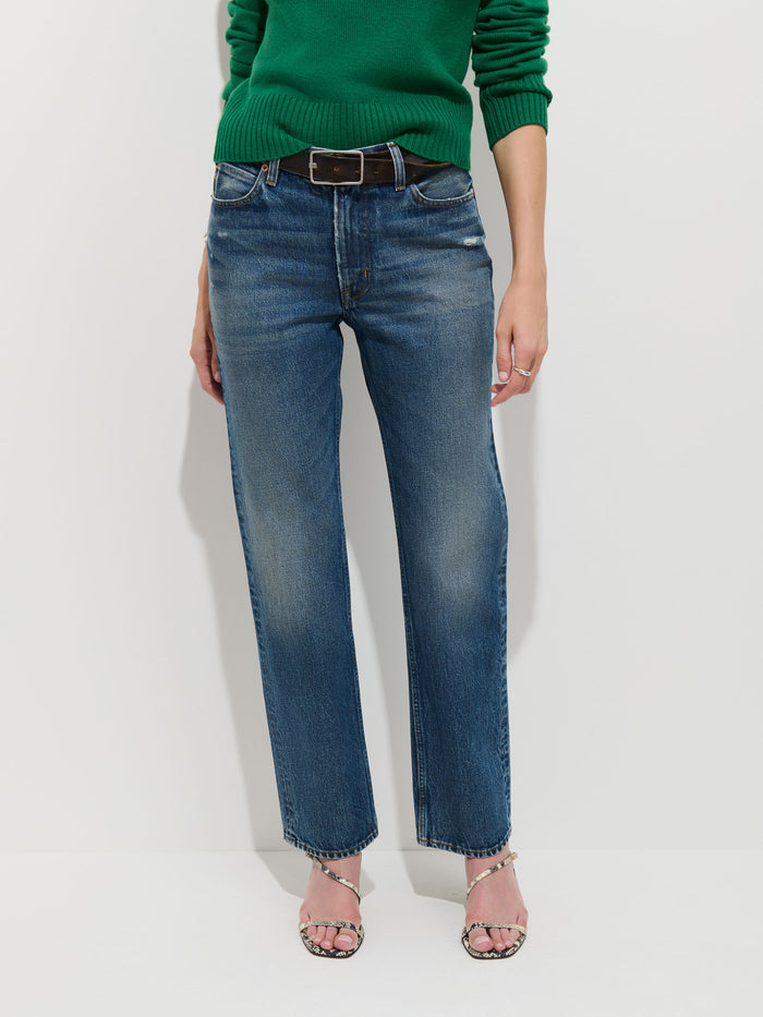 A person stands against a white background, dressed in a green sweater, the Jane Mid-Rise Straight Jean—vintage straight-leg blue jeans made from 100% cotton denim—a black belt, and open-toed heeled sandals. Their face is not visible.