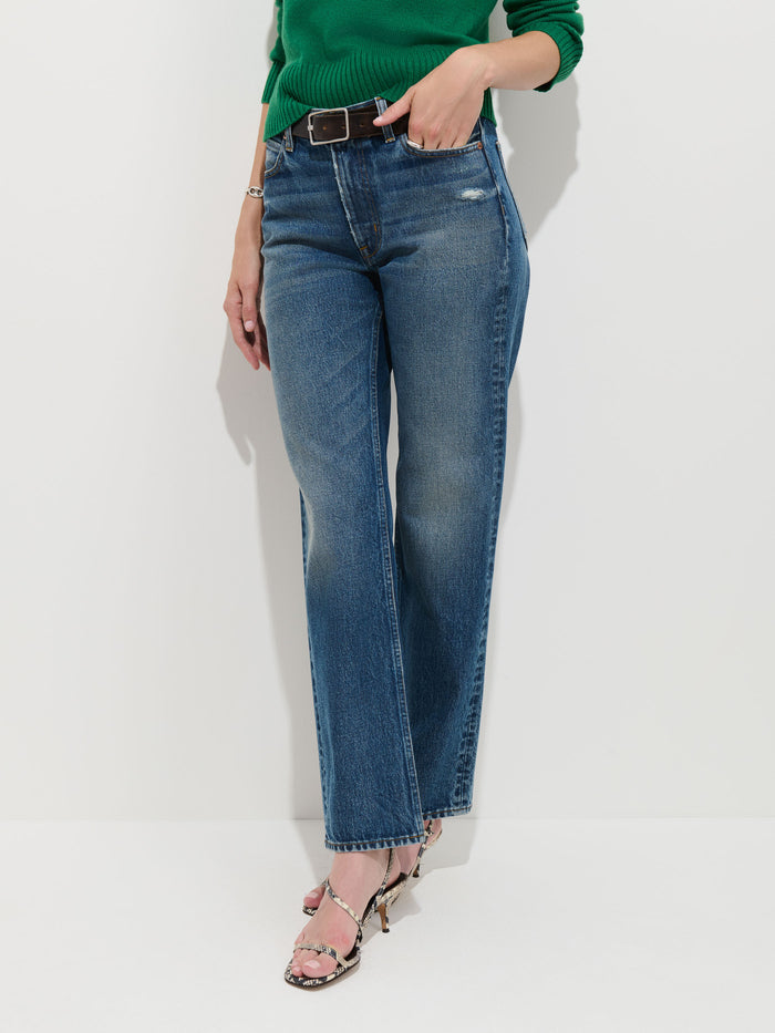 A person wearing the Jane Mid-Rise Straight Jean, paired with a green sweater, a black belt, and snakeskin heels, stands against a white background.