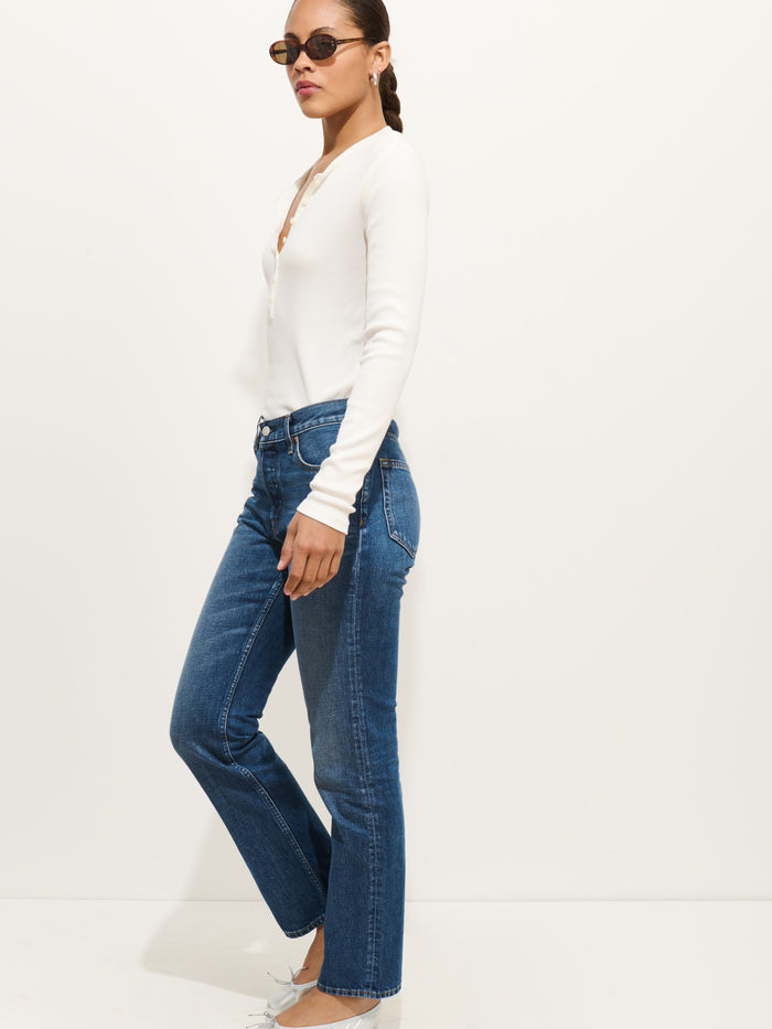 A woman in side profile poses against a white wall, wearing the Jane Mid Rise Straight Jean, a white long-sleeve top, sunglasses, and light-colored pointed shoes. Her hair is braided.