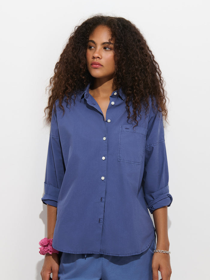 A woman with curly hair wears the Jo Shirt in Cotton Poplin, a loose 100% cotton blue button-up featuring French seams and rolled sleeves, paired with blue pants, a pink scrunchie on her wrist, and a silver bracelet, standing against a plain white background.