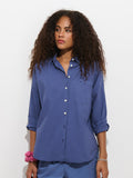A woman with curly hair wears the Jo Shirt in Cotton Poplin, a loose 100% cotton blue button-up featuring French seams and rolled sleeves, paired with blue pants, a pink scrunchie on her wrist, and a silver bracelet, standing against a plain white background.