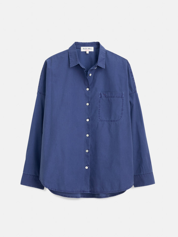 The Jo Shirt In Cotton Poplin is a long-sleeved, button-up blue shirt made from 100% cotton, featuring a single chest pocket and refined French seams, showcased against a plain white background.