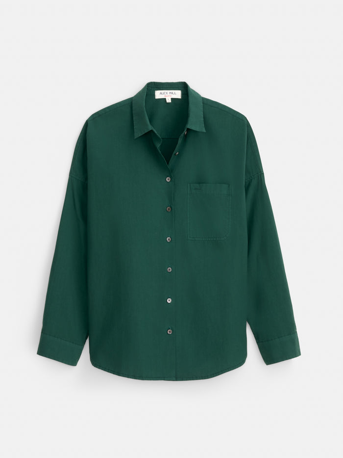 The Jo Shirt In Cotton Poplin is a dark green, long-sleeved button-up shirt made from 100% cotton, featuring a single chest pocket and elegant Troca shell buttons, displayed against a plain white background.