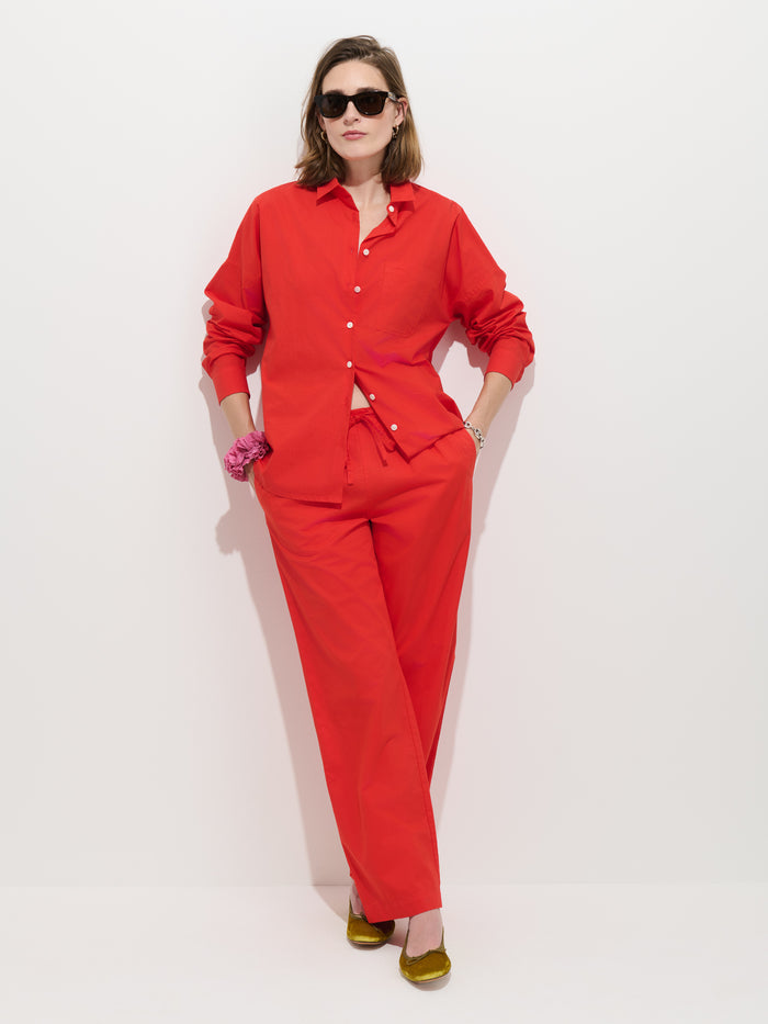 Wearing the Jo Shirt in Cotton Poplin—a bright red, 100% cotton button-up with Troca shell buttons—paired with matching red pants, this person stands against a white background. With hands in their pockets, they accessorize with sunglasses, gold shoes, and a pink scrunchie on their wrist.