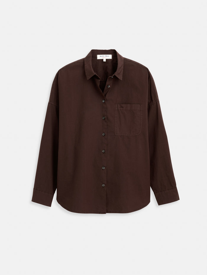 The Jo Shirt In Cotton Poplin is a dark brown, long-sleeve button-up made from 100% cotton, featuring a single chest pocket and Troca shell buttons, shown against a white background.