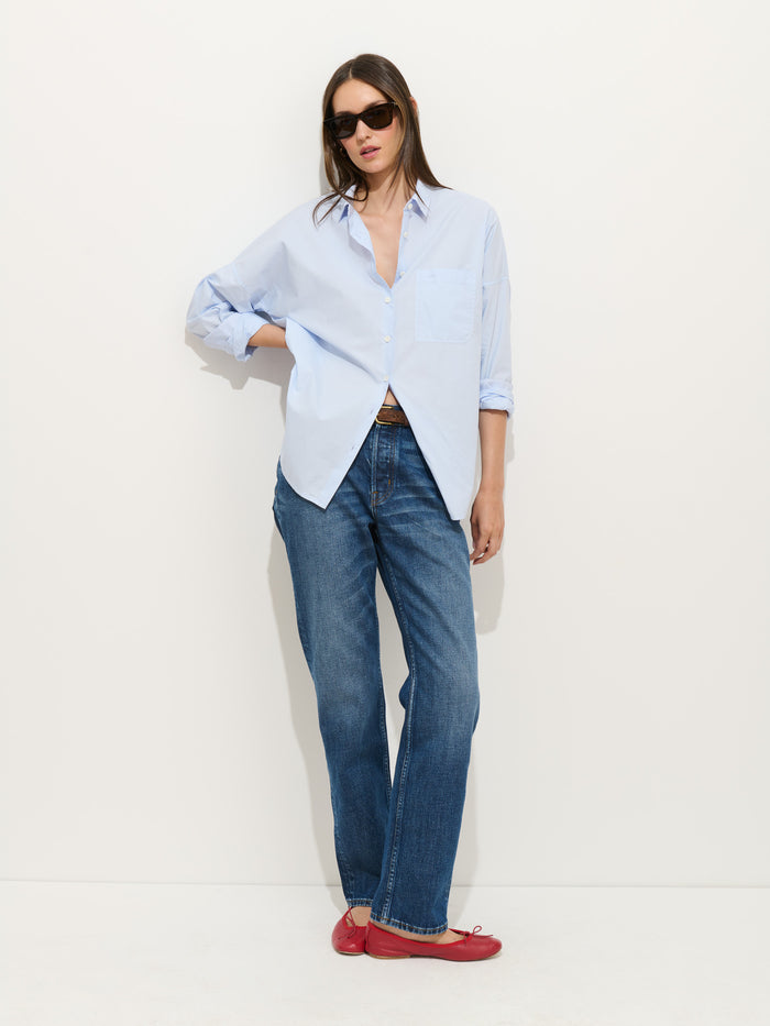 A woman in the Jo Shirt In Cotton Poplin, styled loosely over blue jeans and red flats, stands against a white wall. She has long brown hair, wears black sunglasses, and has one hand tucked in her pocket.