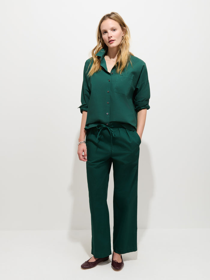 A woman stands against a plain white background, wearing the Jo Shirt in Cotton Poplin—a loose-fitting, dark green 100% cotton button-up with Troca shell buttons—and matching trousers. She pairs the outfit with brown flat shoes. Her hands are relaxed at her sides and her straight, shoulder-length hair frames her face.