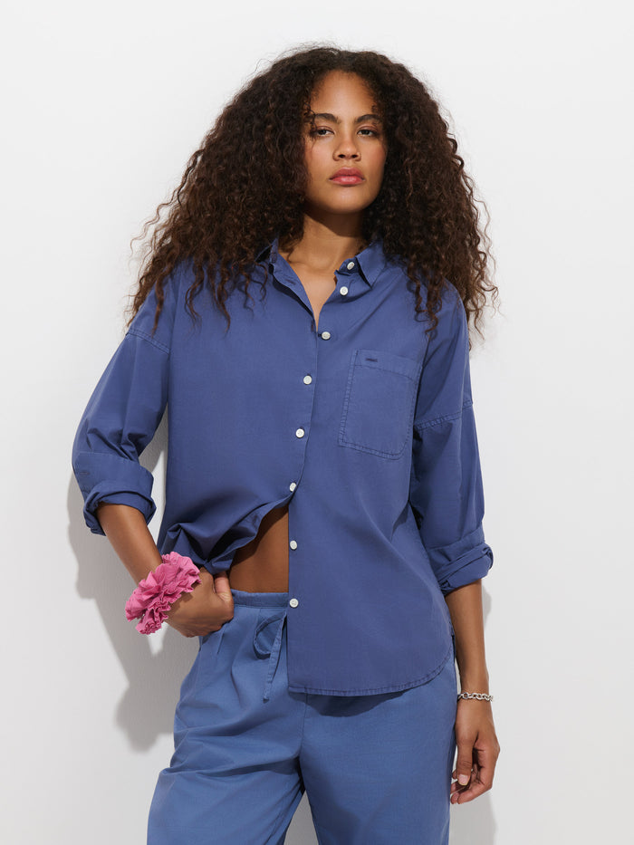 A woman with curly hair stands against a white background, wearing the Jo Shirt in cotton poplin—a loose blue 100% cotton button-up—paired with matching blue pants. She has a pink scrunchie on her wrist and a relaxed, confident expression.