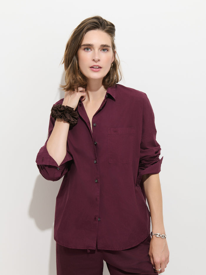 A woman with shoulder-length brown hair wears the Jo Shirt in Cotton Poplin in maroon, paired with matching pants, standing against a plain white background. She rests one hand near her neck and looks directly at the camera.
