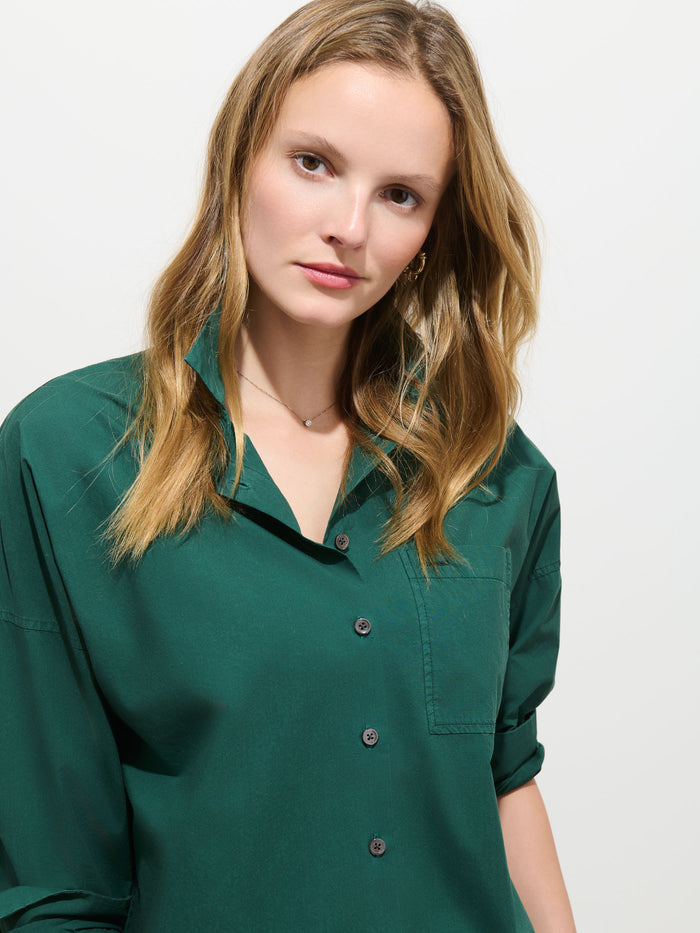 A person with long, light brown hair wears the Jo Shirt In Cotton Poplin—a green 100% cotton button-up featuring Troca shell buttons—and looks at the camera with a neutral expression against a plain white background.