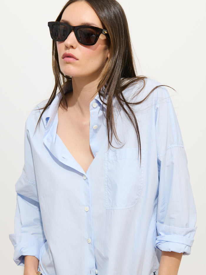 A woman with straight brown hair wears black sunglasses and the Jo Shirt In Cotton Poplin, a light blue oversized style with rolled-up sleeves, standing against a plain light background.