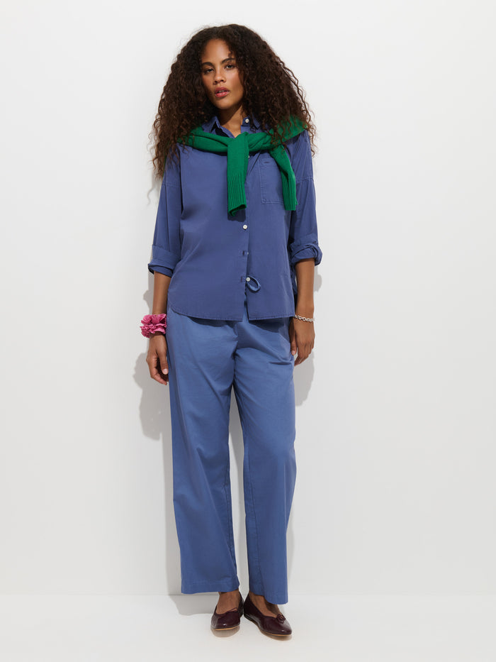 A woman with curly hair wears the Jo Shirt in Cotton Poplin, a blue 100% cotton button-up featuring French seams, paired with blue pants, a green sweater draped over her shoulders, maroon shoes, and pink bracelets, as she stands against a white background.