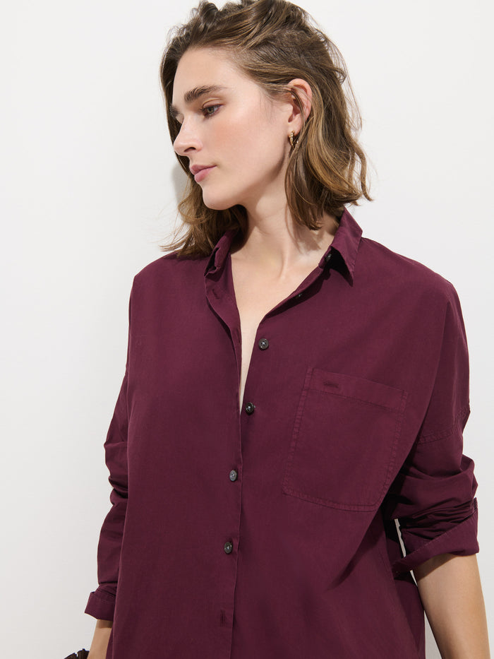 Woman with shoulder-length brown hair wearing the Jo Shirt in Cotton Poplin—a loose, long-sleeve maroon button-up crafted from 100% cotton and finished with Troca shell buttons—standing against a plain white background, looking to the side with a neutral expression.