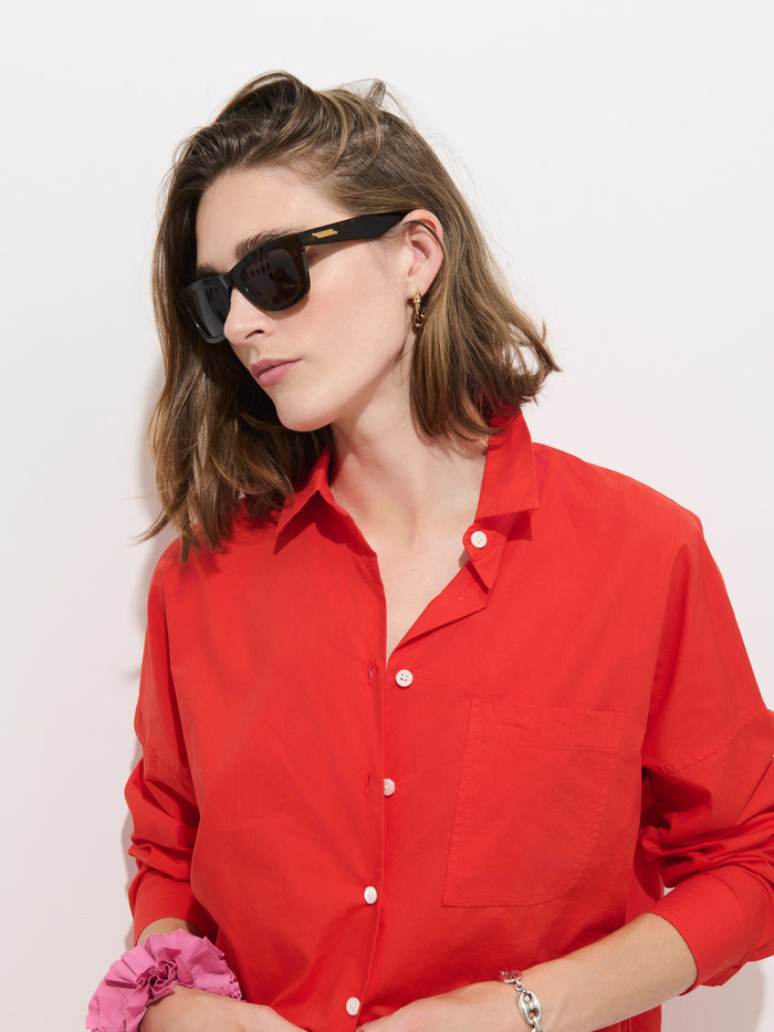 A person wearing dark sunglasses and the Jo Shirt In Cotton Poplin—a bright red, 100% cotton button-up shirt with Troca shell buttons—stands against a plain white background, holding a pink scrunchie in one hand.