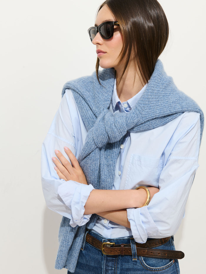 A woman in sunglasses wears the Jo Shirt in Cotton Poplin in light blue with jeans, a brown belt, a blue knit sweater draped over her shoulders and tied in front, plus a gold bracelet. Her arms are crossed as she poses.