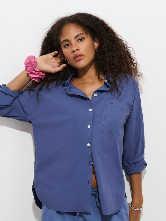 A woman with curly hair, wearing the Jo Shirt in Cotton Poplin—a blue, 100% cotton button-up shirt with French seams—and a pink scrunchie on her wrist stands against a plain white background, resting one hand near her ear.